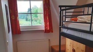 A dormitory room with bunk beds, a window with red curtains, a radiator, and a wooden desk. - Apartment 1 / o. l -. Rügen-Fewo 47 (Putbus)