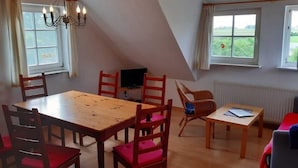 A dining area with a wooden table and chairs, a chandelier, a television, and windows with curtains. - Apartment 1 / o. l -. Rügen-Fewo 47 (Putbus)