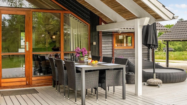A wooden deck with a table and chairs, a wicker sofa, and a stone planter. - Amazing home in Glesborg with sauna (Glesborg)