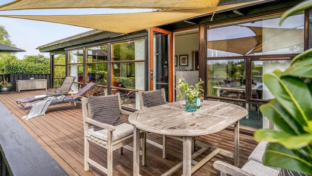 Une terrasse extérieure aménagée avec du mobilier en bois, une table et des chaises, entourée de grandes fenêtres et dotée d’un espace ombragé.