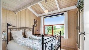 Une chambre avec un plafond en bois, un lit avec un cadre en métal, une fenêtre avec des rideaux et une vue sur la verdure à l’extérieur.