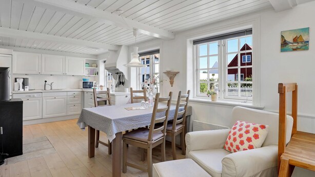 A bright kitchen with white cabinets, a dining table with chairs, and a view of a red house through a window. - 1 bedroom gorgeous apartment in Svaneke (Svaneke)