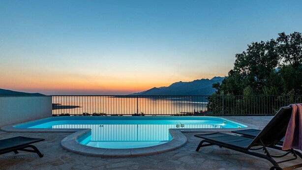 Piscina con vista al atardecer, sillas de descanso y terraza de piedra.