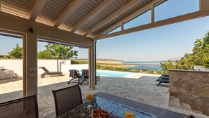Un área de comedor al aire libre con una mesa lista para dos personas, con vistas a una piscina y un paisaje marino.