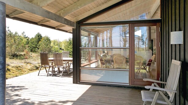 A covered outdoor area with a wooden deck, a glass door leading to an interior space with wicker furniture, and a view of trees and sky. - Gorgeous home in ålbæk with sauna (Ålbæk)