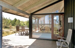 A covered outdoor area with a wooden deck, a glass door leading to an interior space with wicker furniture, and a view of trees and sky. - Gorgeous home in ålbæk with sauna (Ålbæk)