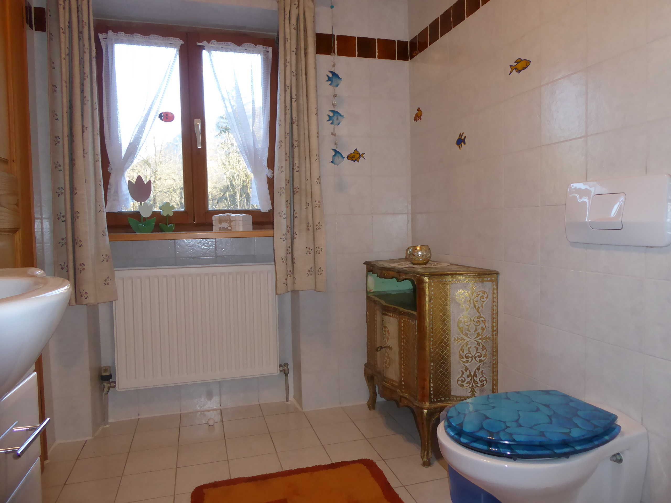 Une salle de bain comprenant des toilettes, un radiateur, une fenêtre avec des rideaux, un meuble décoratif et un lavabo recouvert de carreaux bleus.