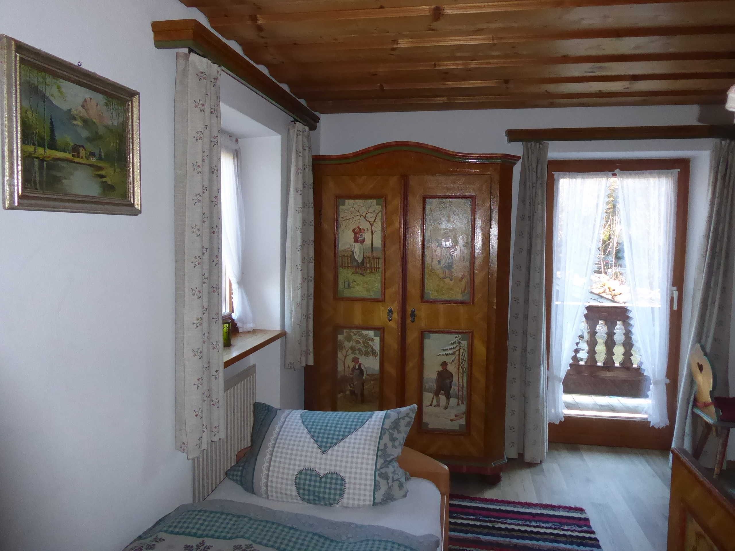 Une chambre avec un plafond en bois, un lit avec une literie à motifs, une armoire en bois ornée d’œuvres d’art, une fenêtre avec des rideaux et une vue sur un balcon.