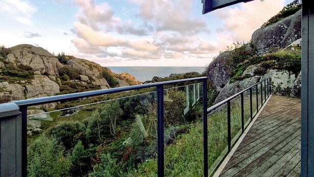Une terrasse en bois, dotée d’une rambarde en métal, qui offre une vue sur une côte rocheuse et la mer.