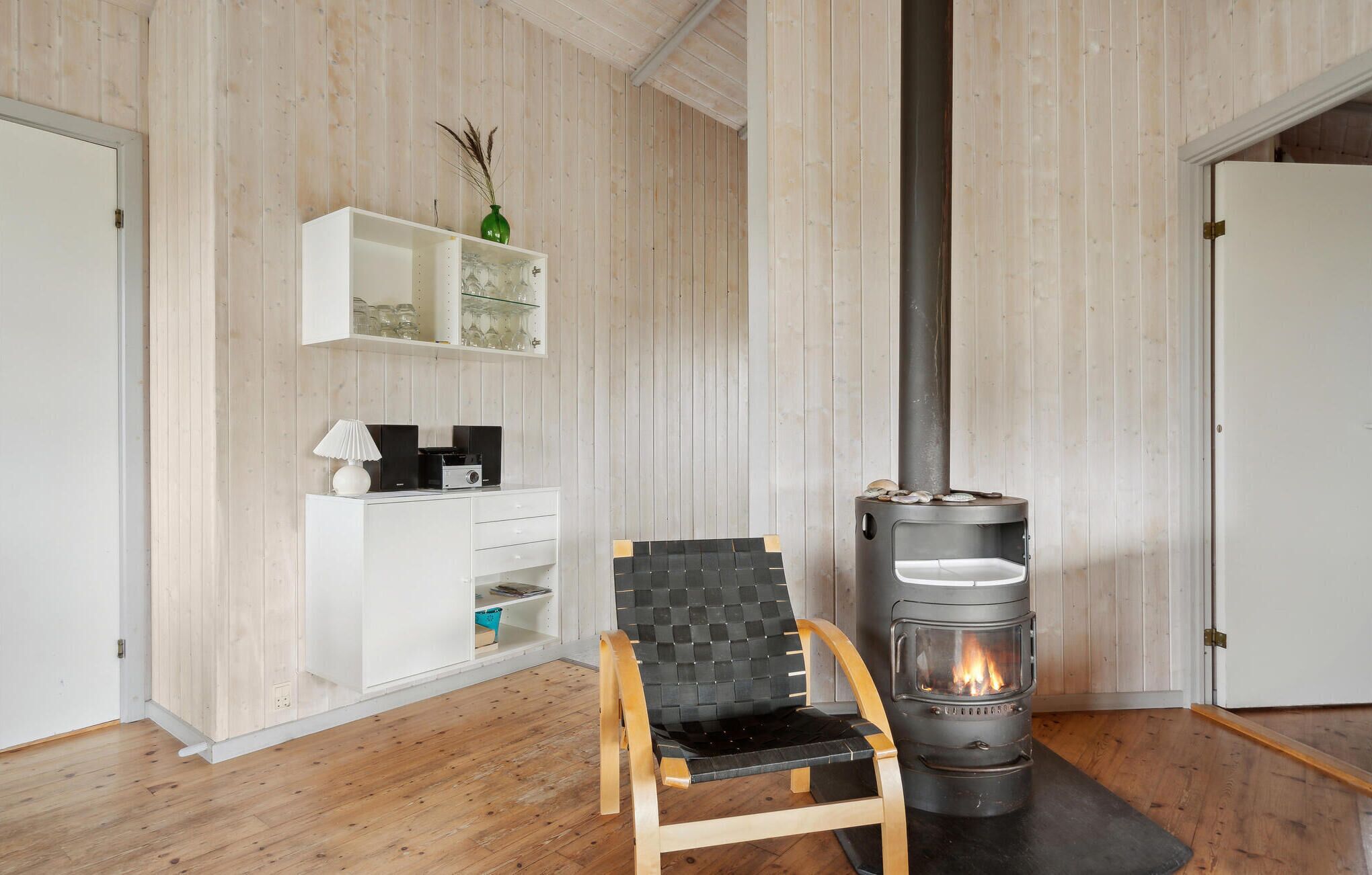 Une pièce avec un plancher en bois, une armoire blanche contenant de la verrerie, une chaise noire et un poêle à bois.