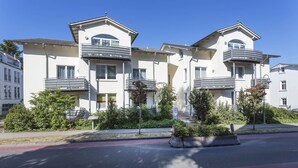 A row of modern, two-story apartment buildings with balconies, surrounded by greenery and a paved street. - Villa a cappella A2 - Villa a cappella (Binz)