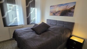 A bedroom with a large bed, a nightstand, a wall-mounted painting, and a window with blinds. - Villa a cappella A2 - Villa a cappella (Binz)