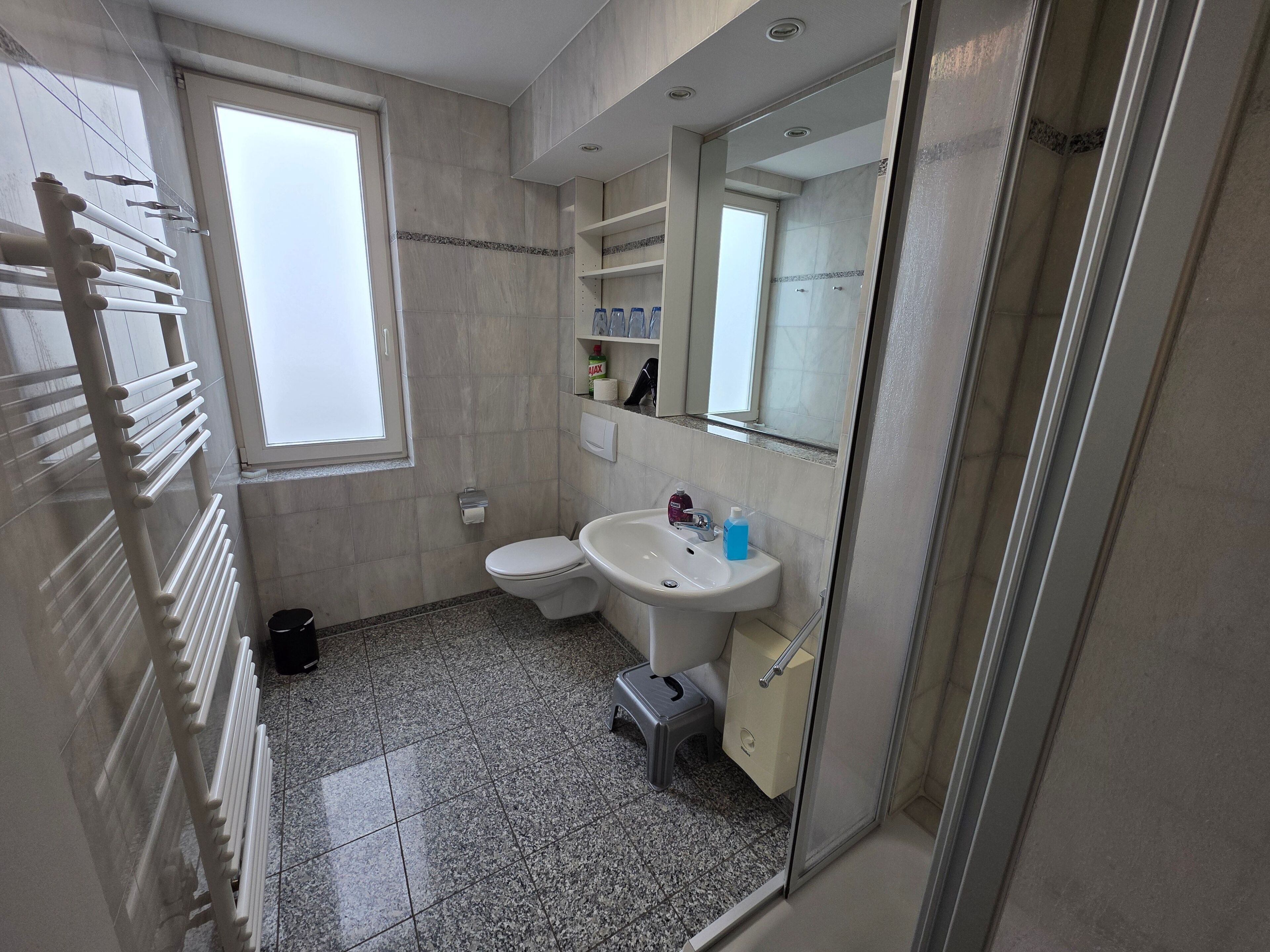 A small bathroom with a toilet, sink, and shower. There is a window providing natural light and a towel rack on the wall.
