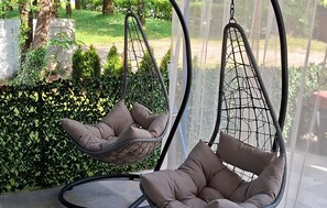 Deux fauteuils suspendus avec coussins, une balustrade en verre et une vue sur un jardin agrémenté d’un mur en pierre et de verdure.