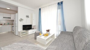 Un saló modern amb un sofà gris, una taula de centre blanca amb begudes, una televisió i una cuina al fons.