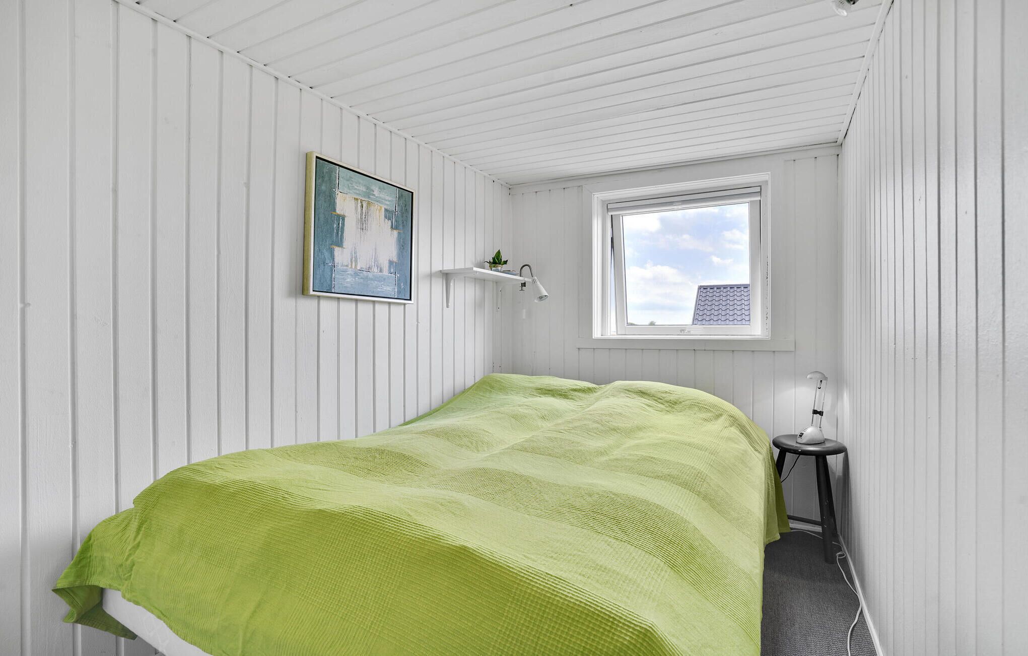 Quarto com colcha verde, um quadro na parede, uma pequena estante com uma planta e uma janela com vista para um edifício.