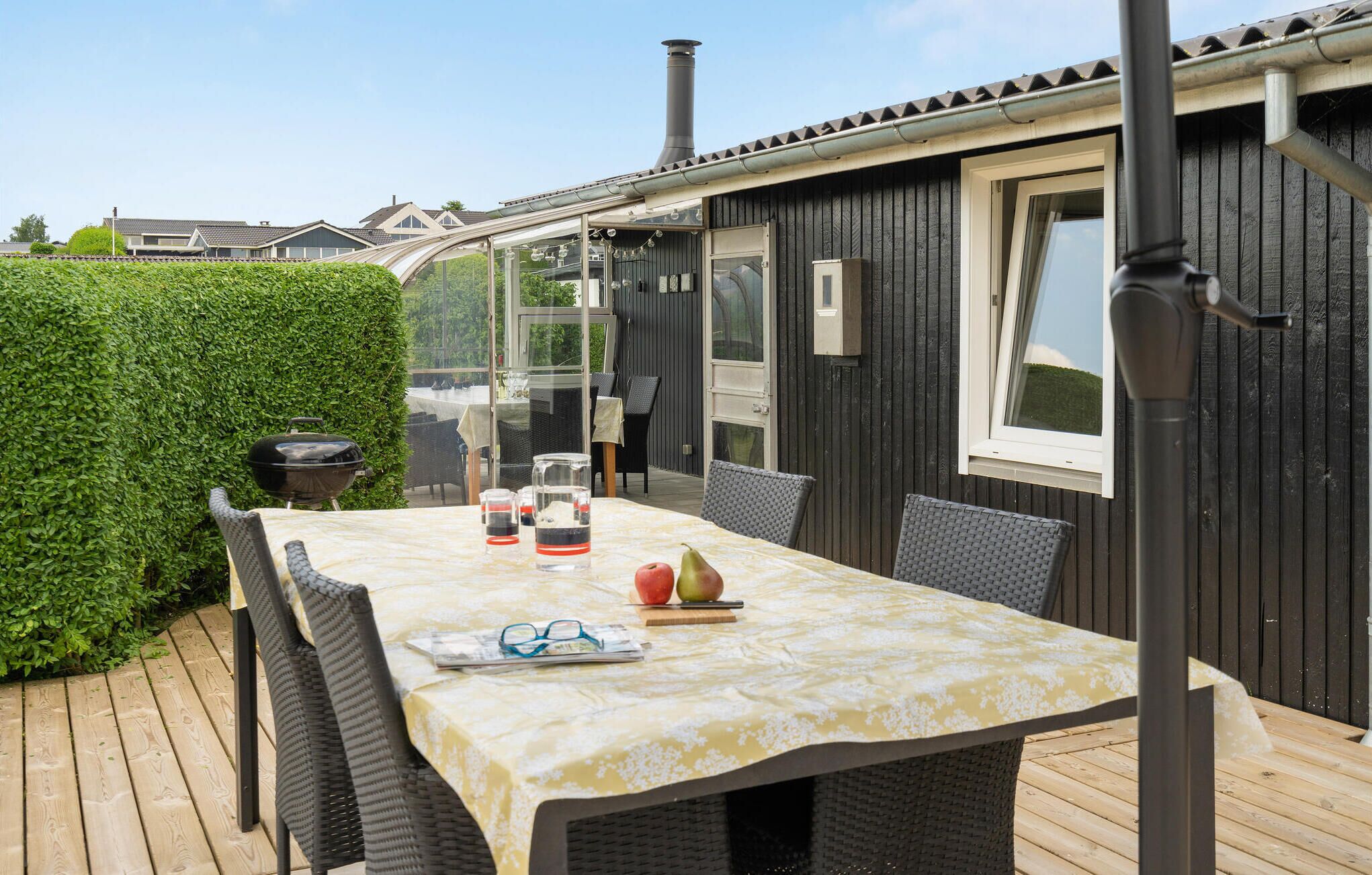 A patio with a table set for two, surrounded by wicker chairs, a grill, and a window overlooking a hedge.