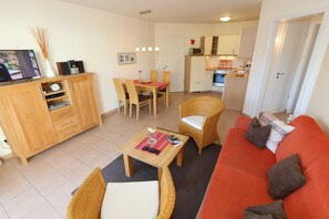 Un salon avec un canapé rouge, des fauteuils en osier, un meuble en bois sur lequel est posée une télévision, et un coin repas avec une table et des chaises.