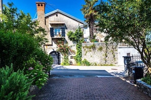A traditional stone building with a tiled roof, a small balcony, and a paved driveway leading to a gate.