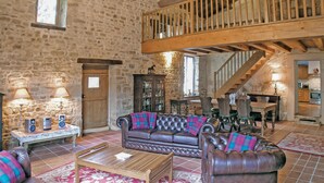 A rustic stone-walled room with a leather sofa, wooden coffee table, and a dining area with a wooden staircase. - Awesome home in Souvigne (Souvigne)