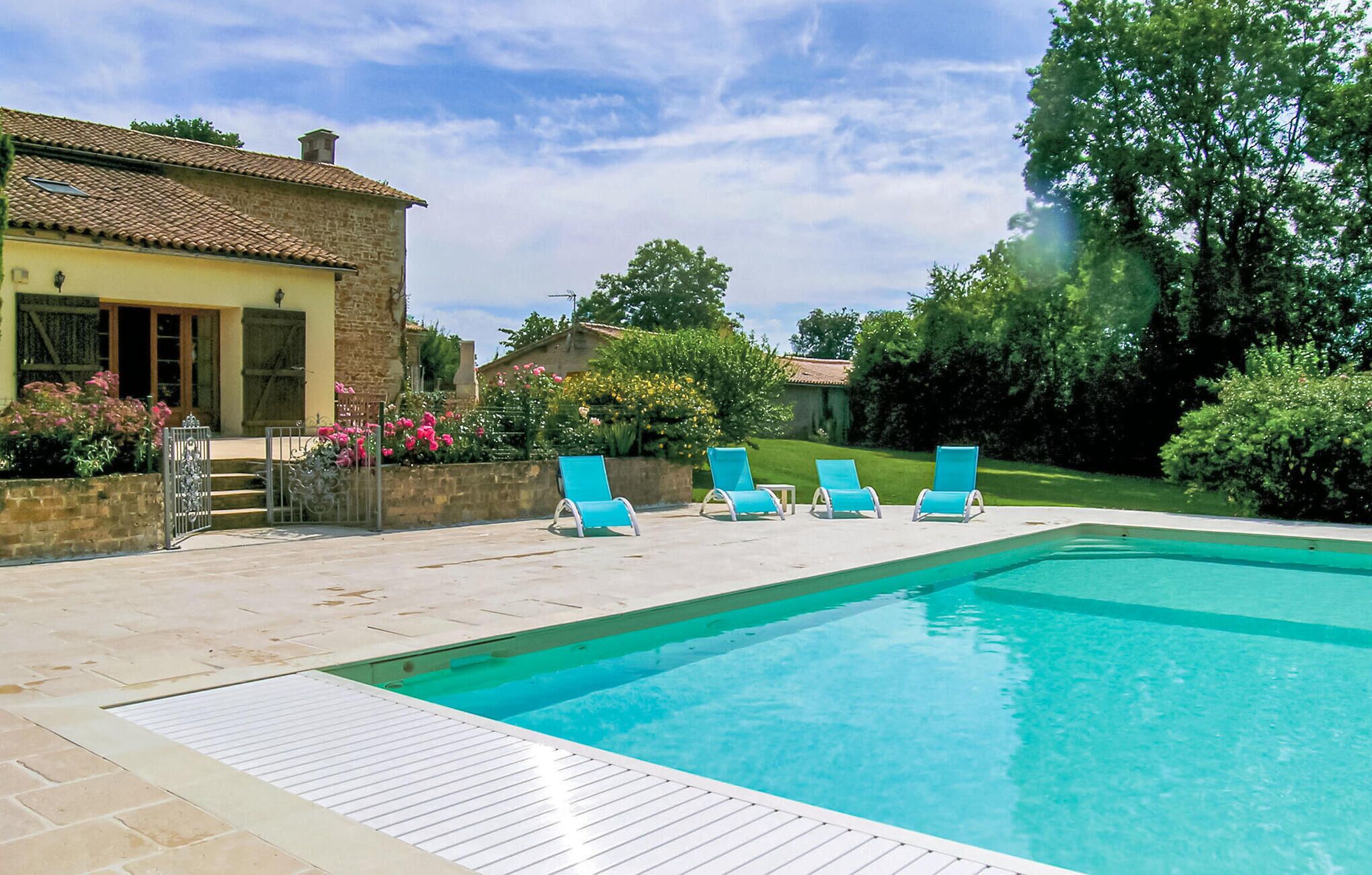 Une piscine entourée de quatre chaises longues bleues, un bâtiment en pierre avec un toit en tuiles et un jardin bien entretenu où poussent diverses plantes et fleurs.
