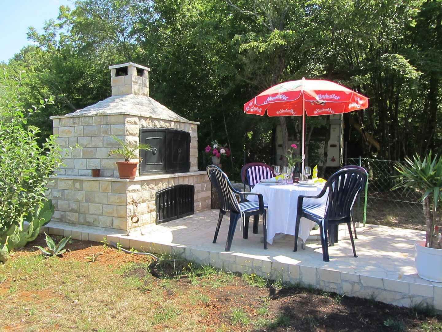 Un patio al aire libre con un horno de pizza de piedra, dos mesas con sillas y un paraguas rojo.