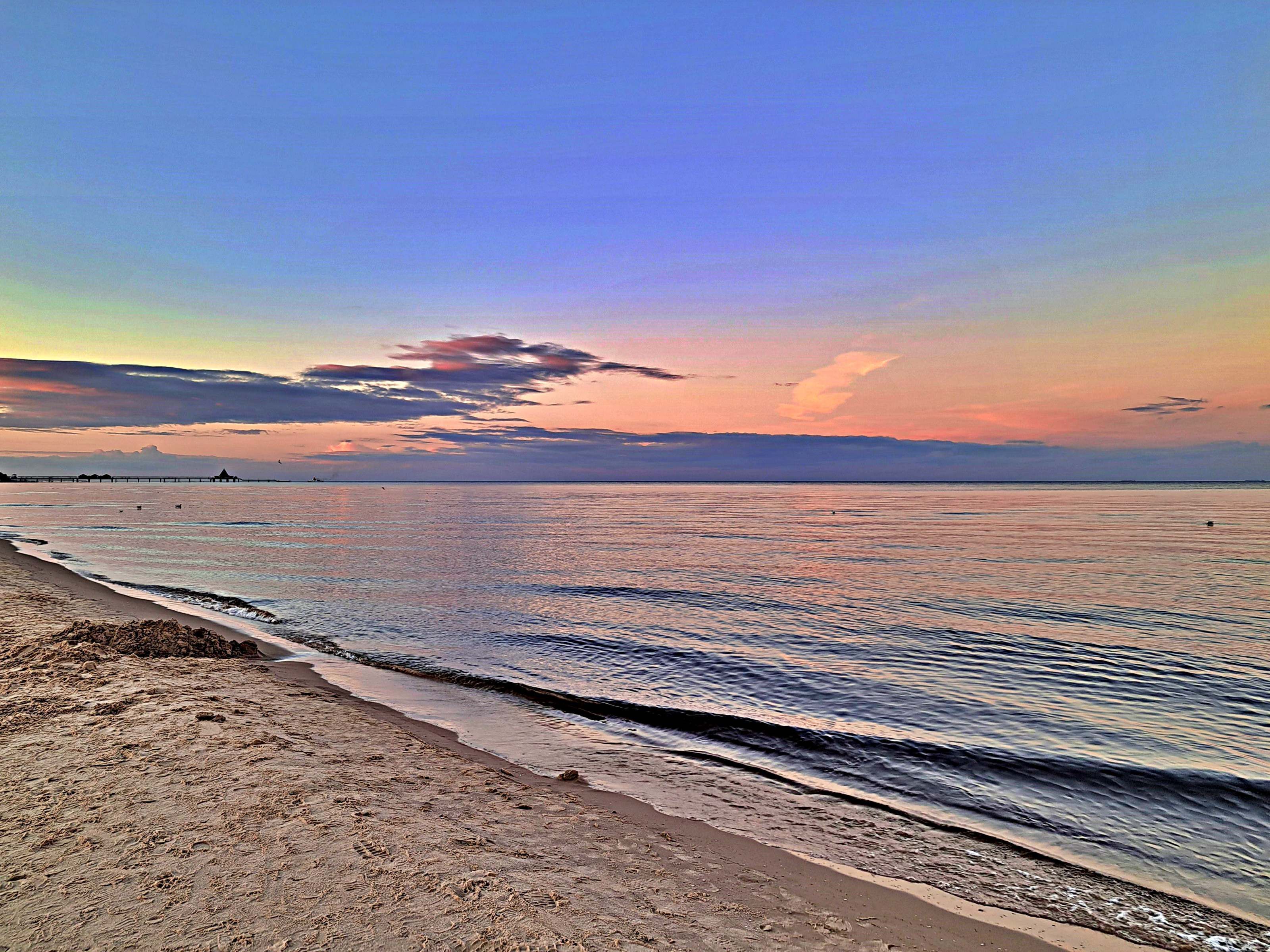 Une plage avec une eau calme, un ciel dégagé et une jetée qui se dessine à l’horizon.