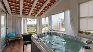 Un bain à remous avec vue sur un jardin et des montagnes, une chaise en osier et une table avec un coussin.