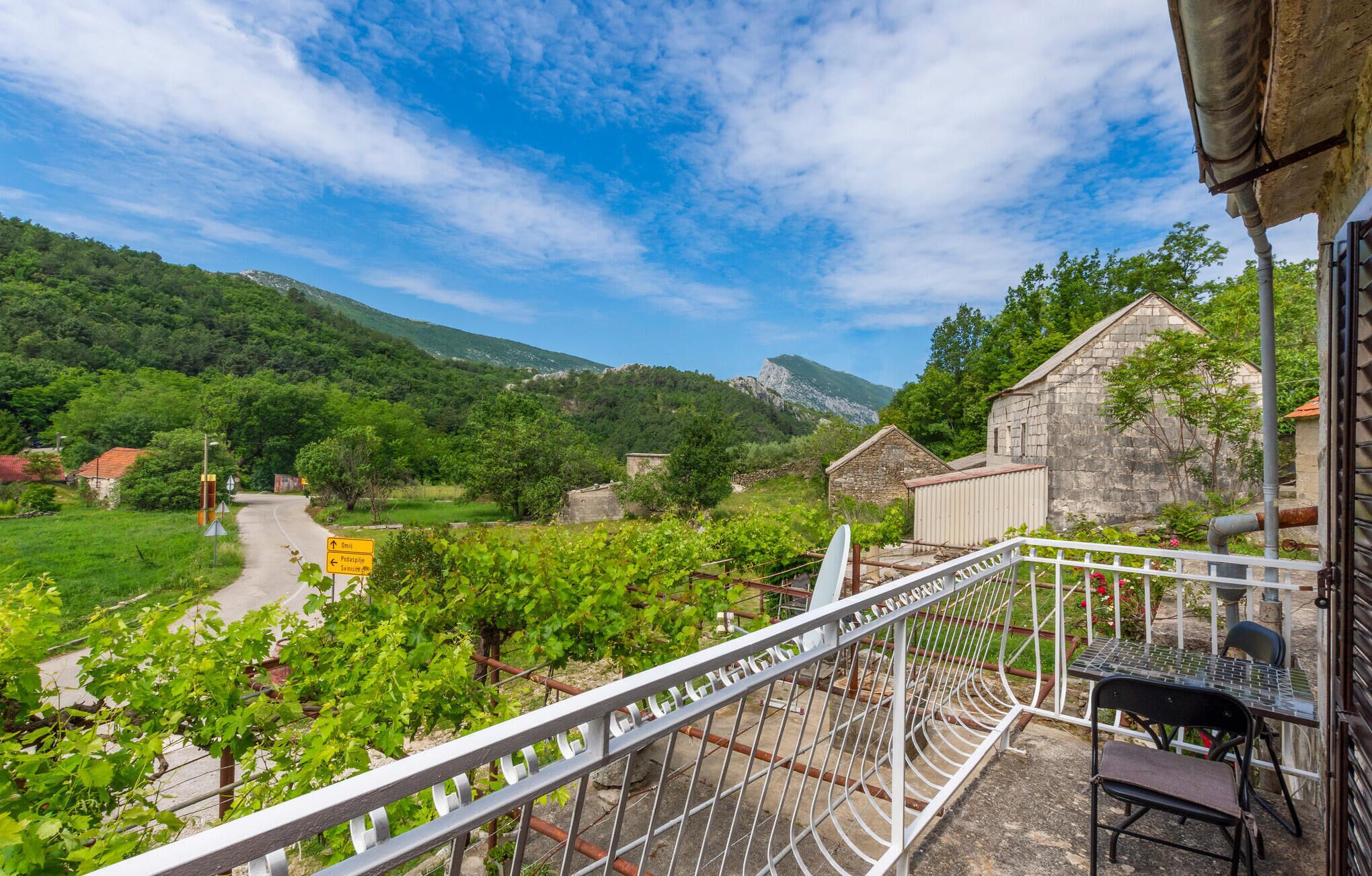 A balcony with a metal railing overlooking a vineyard and a road leading to a mountain.