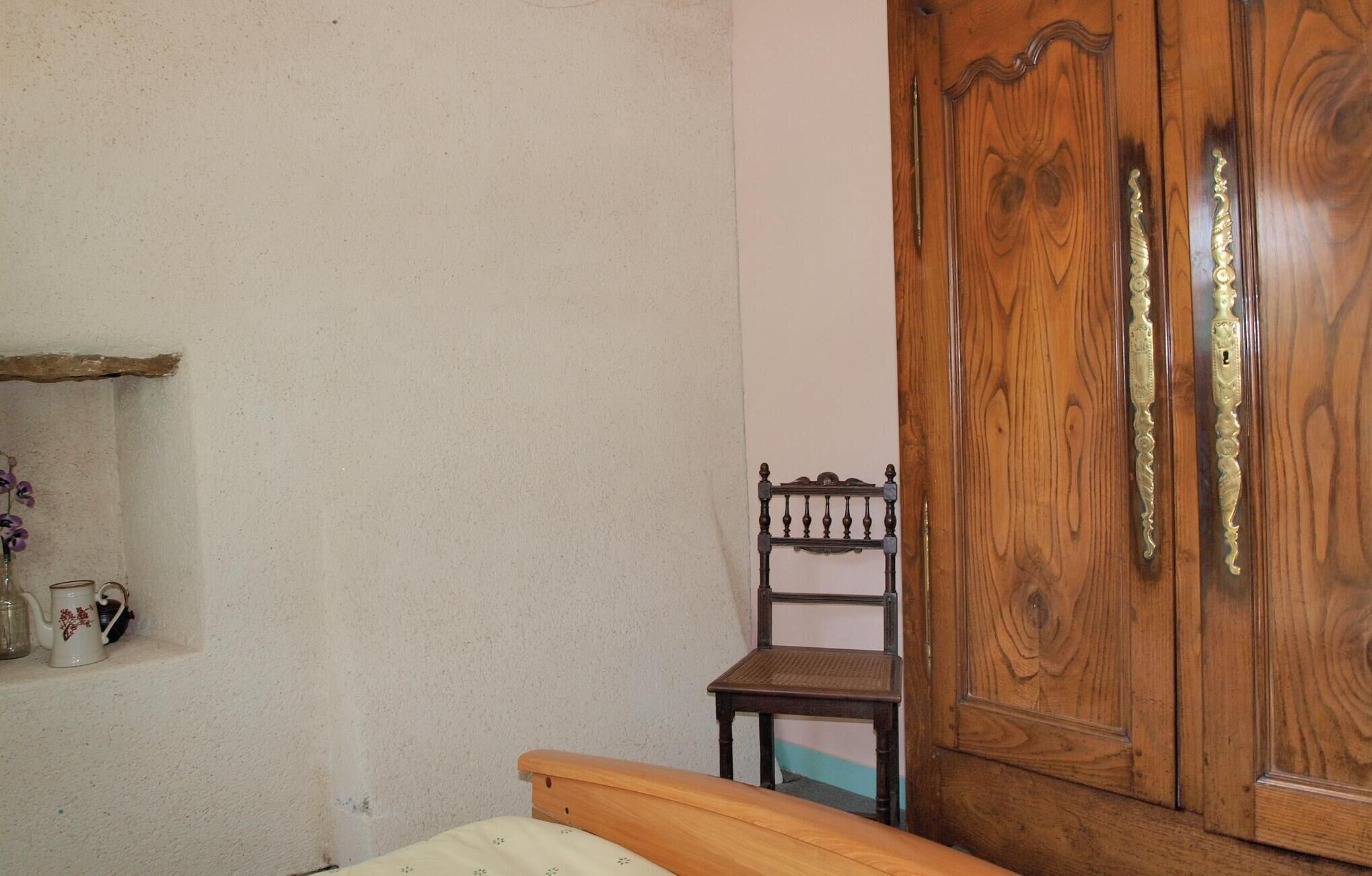 A bedroom with a wooden bed, a small wooden chair, a wooden wardrobe, and a shelf with decorative items.