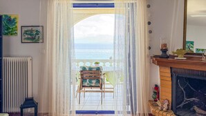 Een kamer met uitzicht op zee door een raam met dunne gordijnen. Er staat een stoel met een tropisch bladmotief, een radiator en een open haard met decoratieve objecten.