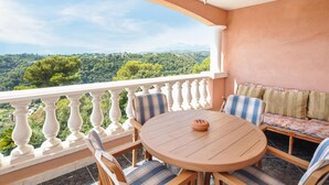 Un balcon avec une table ronde en bois et des chaises, un banc rembourré avec des coussins, et une vue sur un paysage verdoyant.