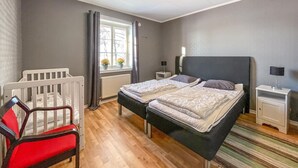 Ein Schlafzimmer mit zwei Betten, einem Babybett, einem Nachttisch mit Lampe und einem Fenster mit Vorhängen.
