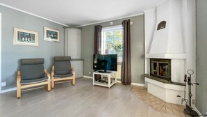Ein Wohnzimmer mit zwei grauen Sesseln, einem Fernseher auf einem weißen Schrank, einem Kamin und einem Fenster mit Vorhängen.