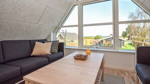 Une pièce moderne avec un plafond en pente, un canapé gris foncé, une table en bois et une grande fenêtre donnant sur l’extérieur.