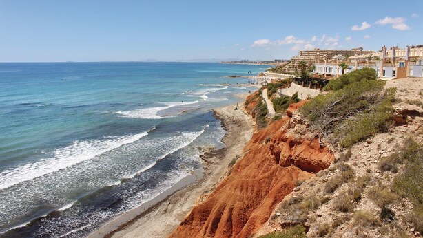 A coastal town with buildings along the cliffside, a beach with waves, and a clear sky.