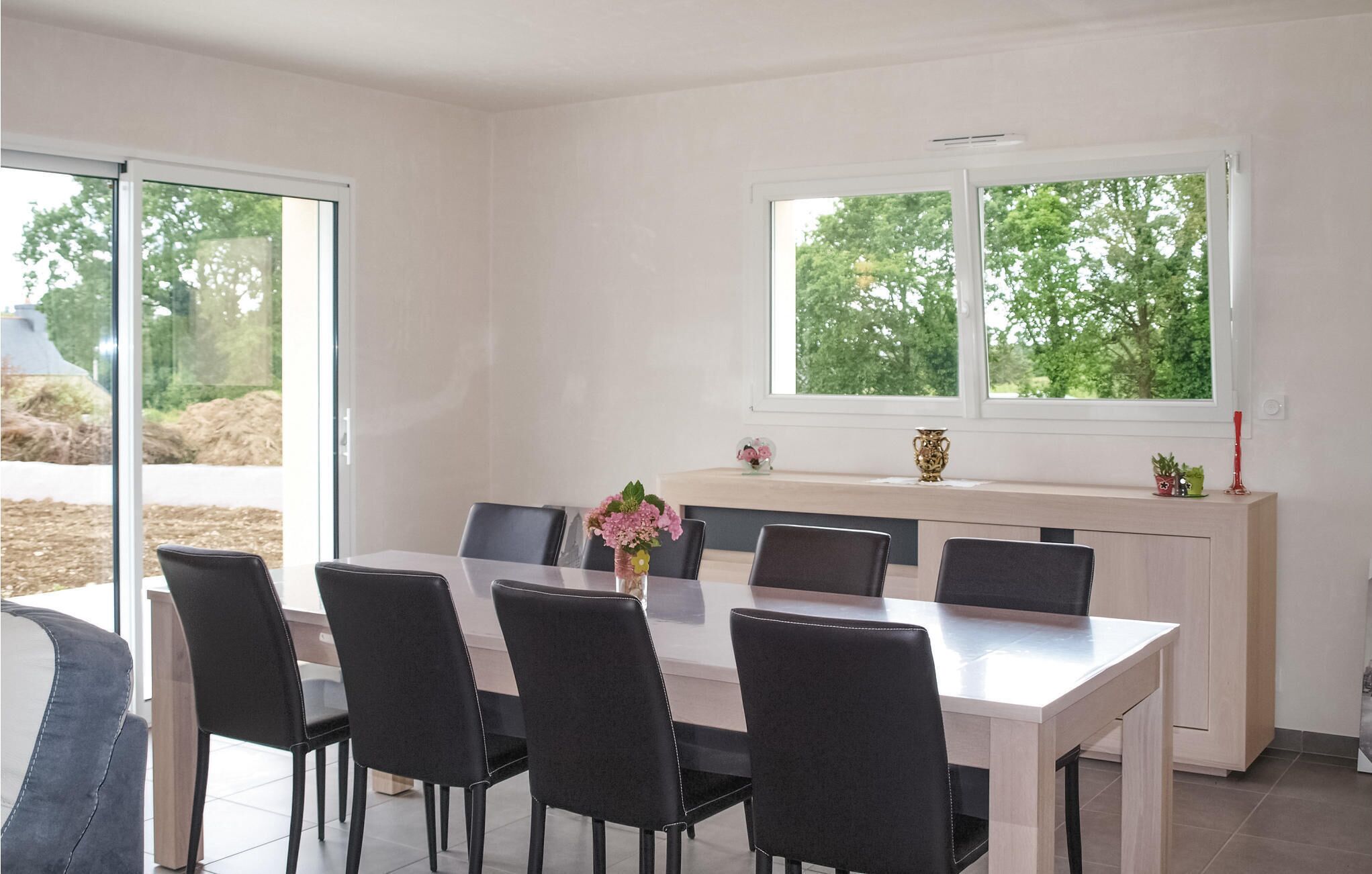 A dining area with a white table and black chairs, a vase with flowers, and a window with a view of trees.