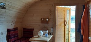 A wooden cabin interior with a bed, a small table, a wall-mounted shelf, and a door leading outside.