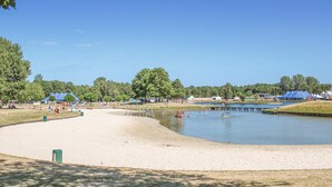 A sandy beach area with a calm water body, surrounded by trees and tents. - Buitengoed Het Lageveld - 65 (Hoge Hexel)