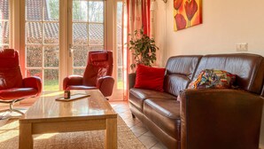 A living room with a brown leather sofa, a red armchair, a wooden coffee table, and a large window with curtains. - Buitengoed Het Lageveld - 65 (Hoge Hexel)