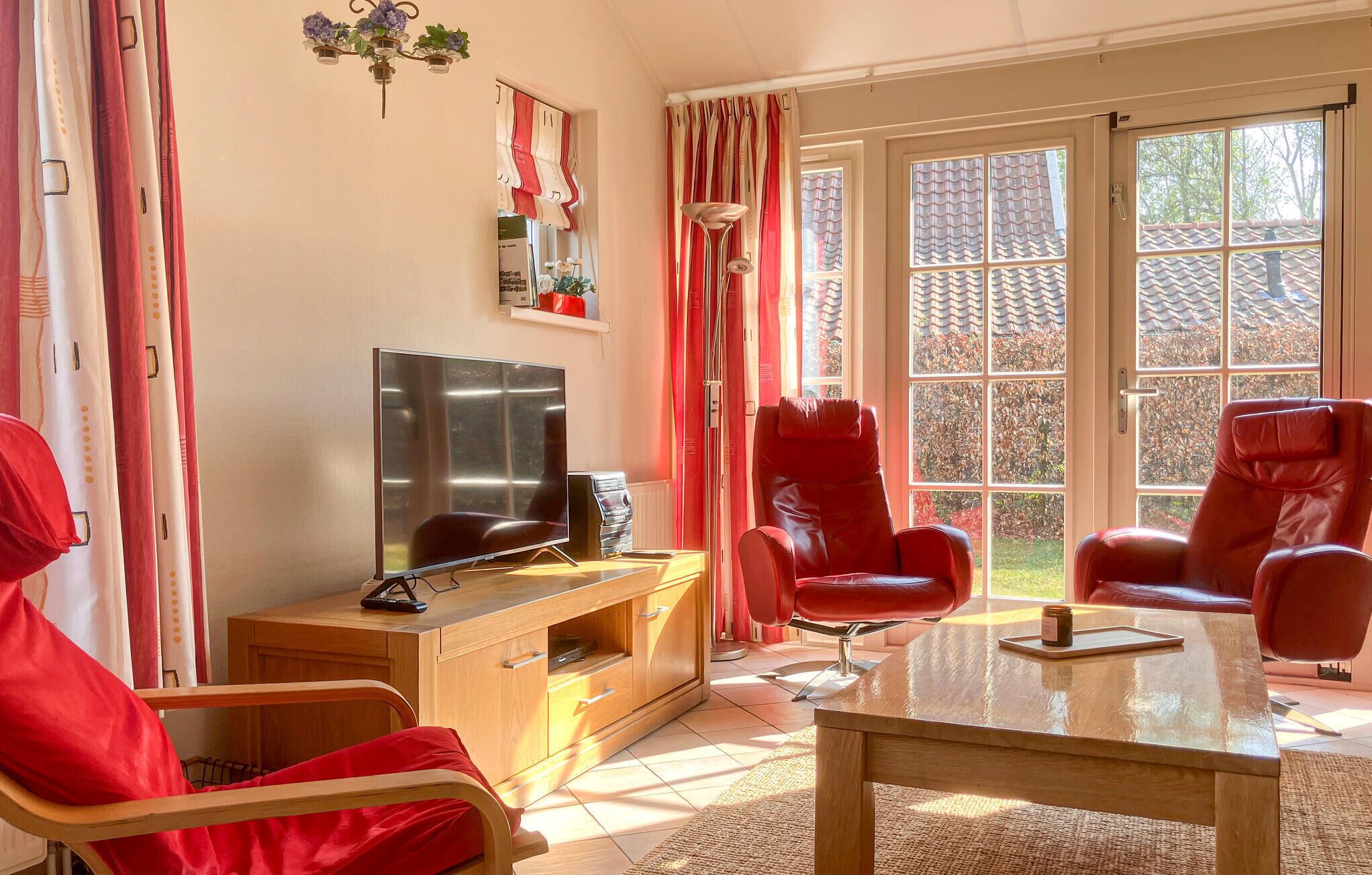 Sala de estar com televisão, móveis de madeira, cadeiras vermelhas e janelas amplas.