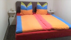 A bed with a colorful bedspread and pillows, a red mattress, and white bedside tables with lamps.