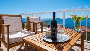 Una mesa de madera con una botella de vino, una copa y un cuenco de cereales en una terraza con vistas al mar.