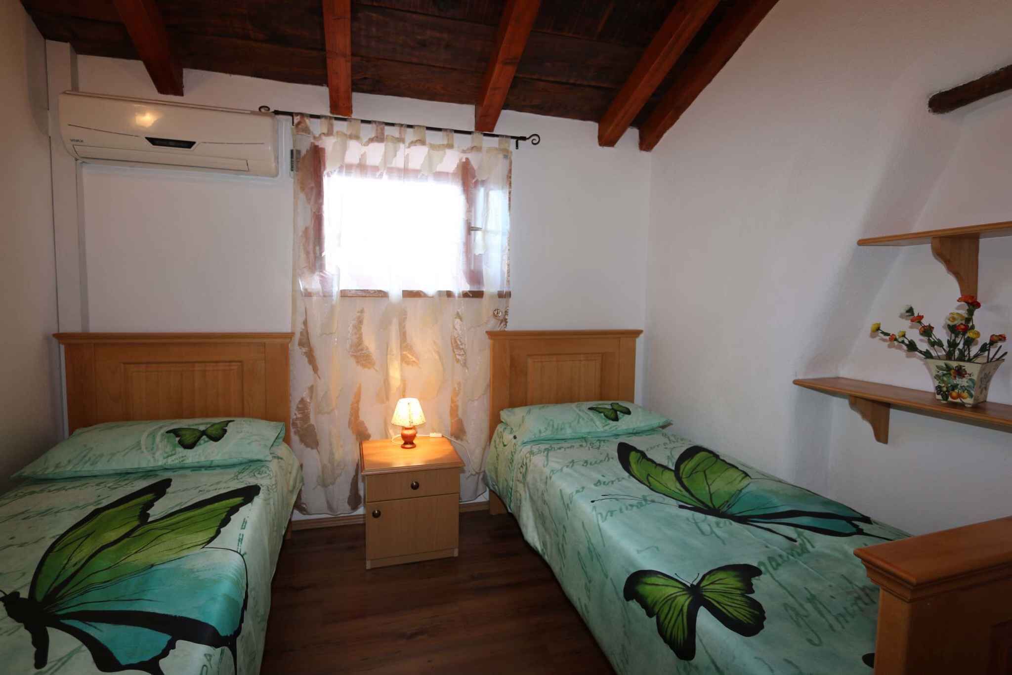 A room with two single beds, wooden headboards, a nightstand with a lamp, a window with sheer curtains, and a small shelf with flowers.