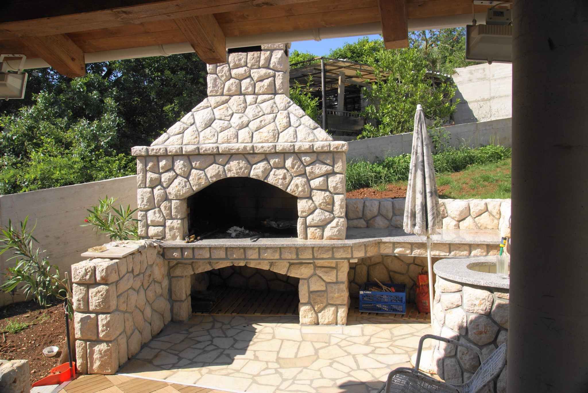 An outdoor stone-built pizza oven with a chimney, surrounded by stone walls and a wooden pergola.