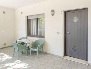 A room with a table and chairs, a window with blinds, a door with a diamond-shaped window, and a wall-mounted light. - Holiday apartment with balcony and air conditioning (Lignano)