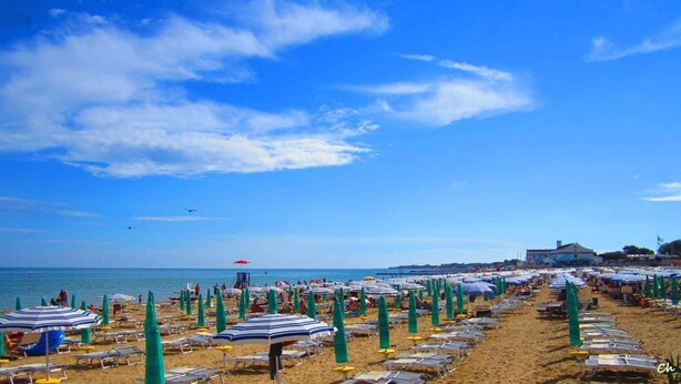A beach with rows of sunbeds and umbrellas, a lifeguard tower, and a clear blue sky. - Holiday apartment with balcony and air conditioning (Lignano)