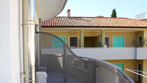 A narrow balcony with a metal pipe, a covered stairway, and a building with a tiled roof in the background. - Holiday apartment with balcony and air conditioning (Lignano)