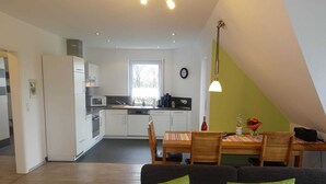 A modern kitchen with white cabinets, a dining area with a wooden table and chairs, and a dark-colored sofa with green cushions. - Ferienwohnung Regenbogenhaus - Ferienwohnung Regenbogenhaus: Special Rate May and June! (Hasselberg)
