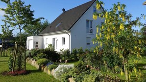 A white two-story house with a sloped roof, surrounded by a well-maintained garden with various plants and flowers. - Ferienwohnung Regenbogenhaus - Ferienwohnung Regenbogenhaus: Special Rate May and June! (Hasselberg)